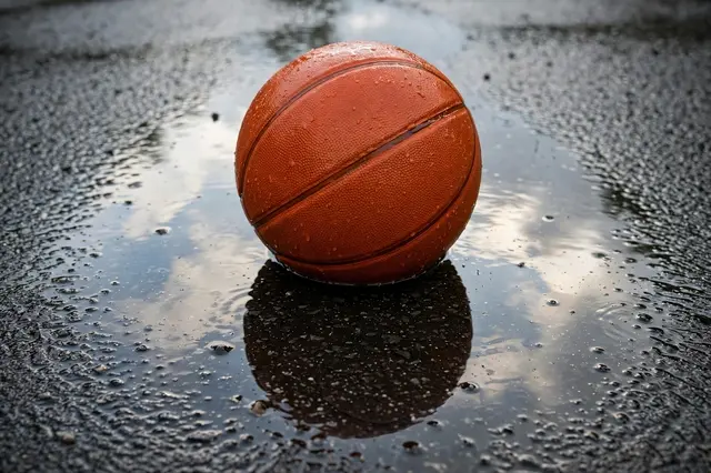 Wet orange basketball sits in a rain puddle on an asphalt court with sky reflections