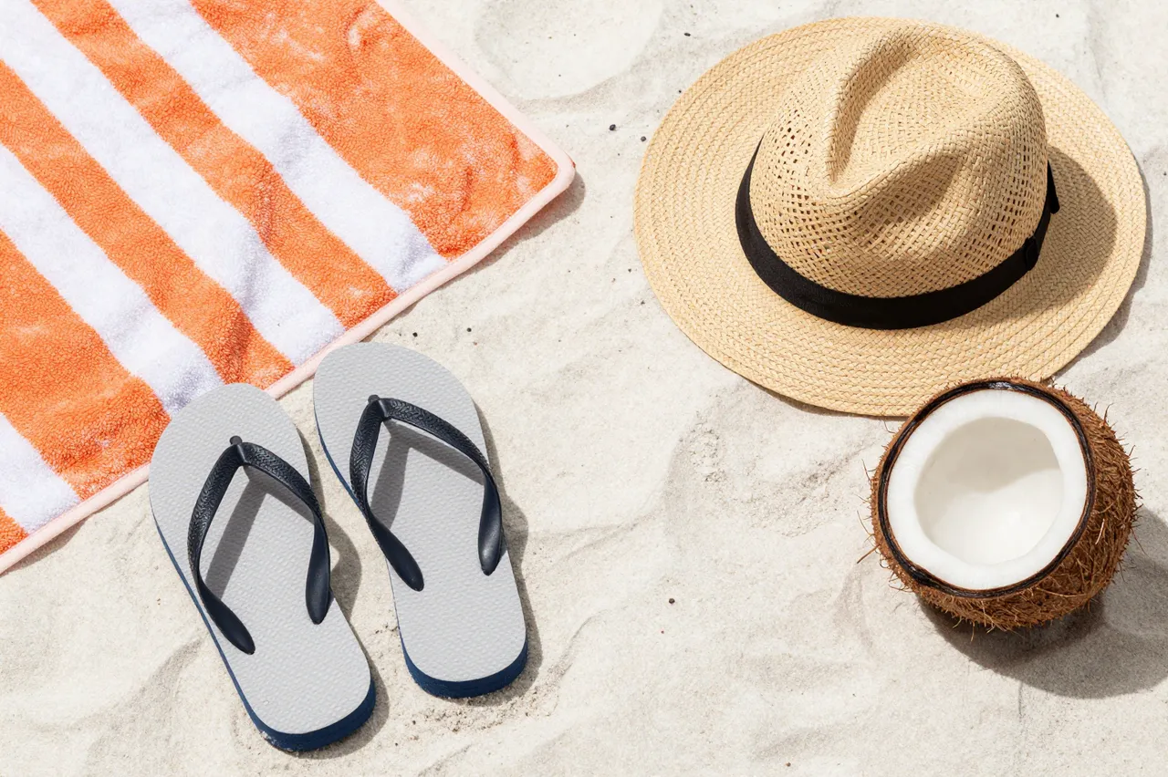 Flat lay of beach towel, straw hat, flip flops and halved coconut arranged on pale beach sand.