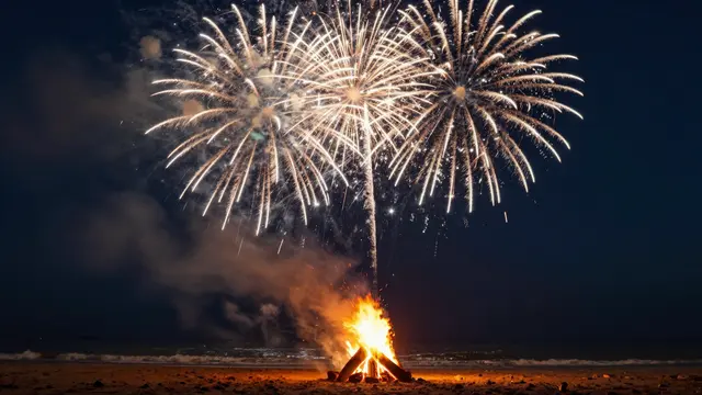 A glowing beach bonfire burns on sandy shore while bright fireworks burst in the dark night sky above the ocean