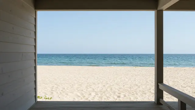 View from a wooden beach house porch looking out over a sandy beach to the calm turquoise ocean under a clear sky