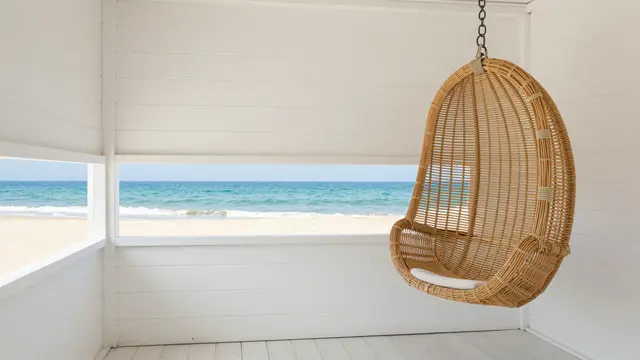 Whitewashed beach hut interior with a hanging rattan chair and sea view through side windows