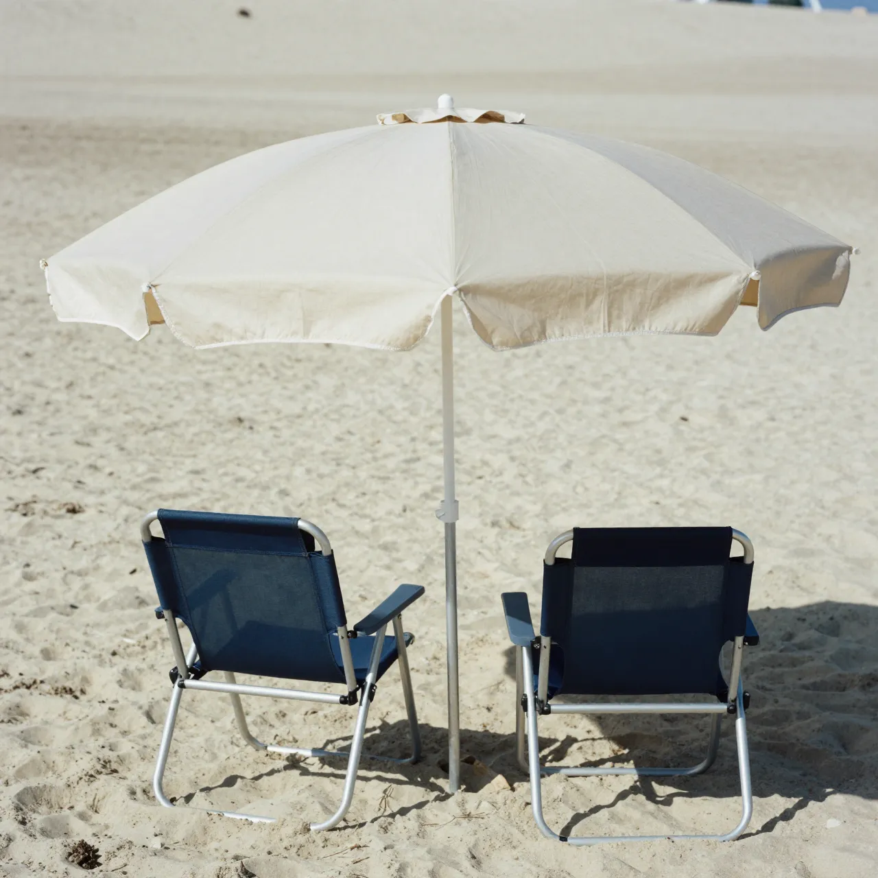 A cream beach umbrella stands between two blue folding chairs on sunlit sand