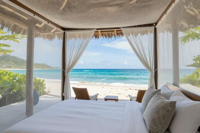 Open-air beach villa bedroom with mosquito netting overlooking a turquoise ocean and sandy shore