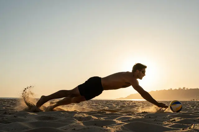 A male beach volleyball player dives across sand toward a volleyball as sand kicks up at golden sunset
