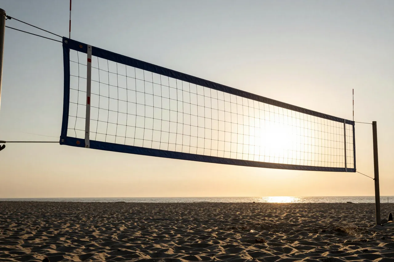 A beach volleyball net stretches across an empty sandy beach with the sun setting low over the ocean horizon.