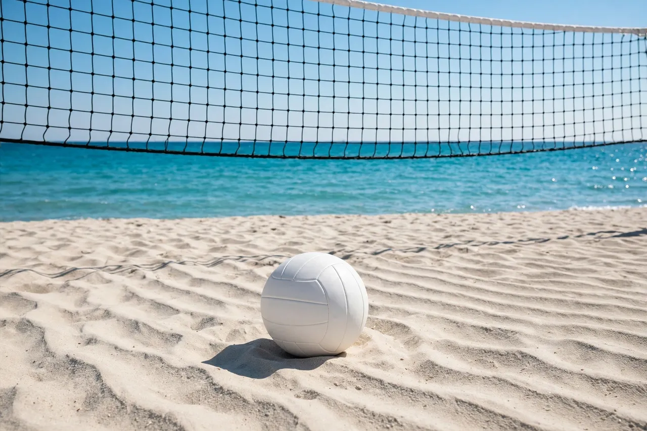 A white volleyball rests on rippled beach sand below a volleyball net with turquoise sea under midday sun.