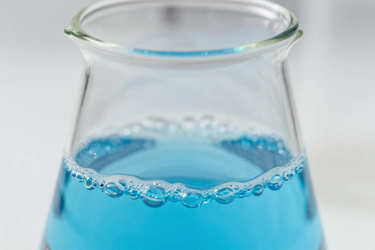 Close-up macro view of a glass beaker holding bubbling bright blue laboratory liquid against a soft light gray background