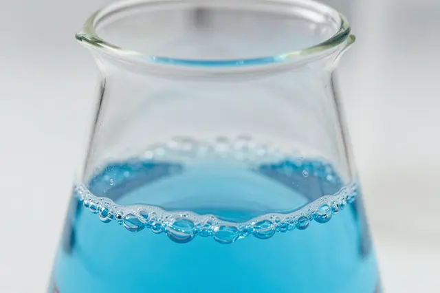 Close-up macro view of a glass beaker holding bubbling bright blue laboratory liquid against a soft light gray background