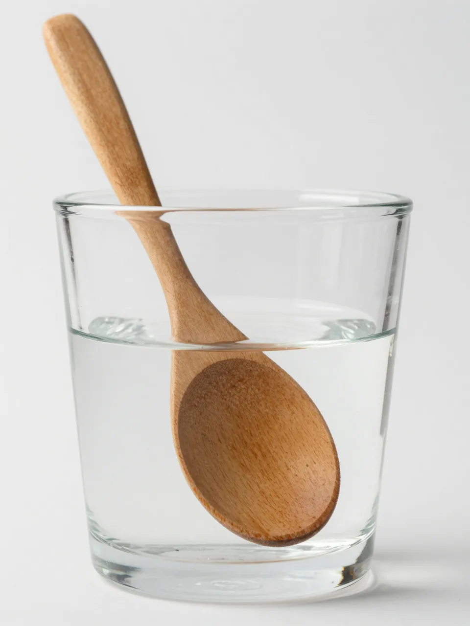 A wooden spoon sits inside a clear glass of water, appearing bent at the water line against a plain white background.