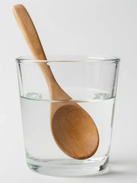 A wooden spoon sits inside a clear glass of water, appearing bent at the water line against a plain white background.