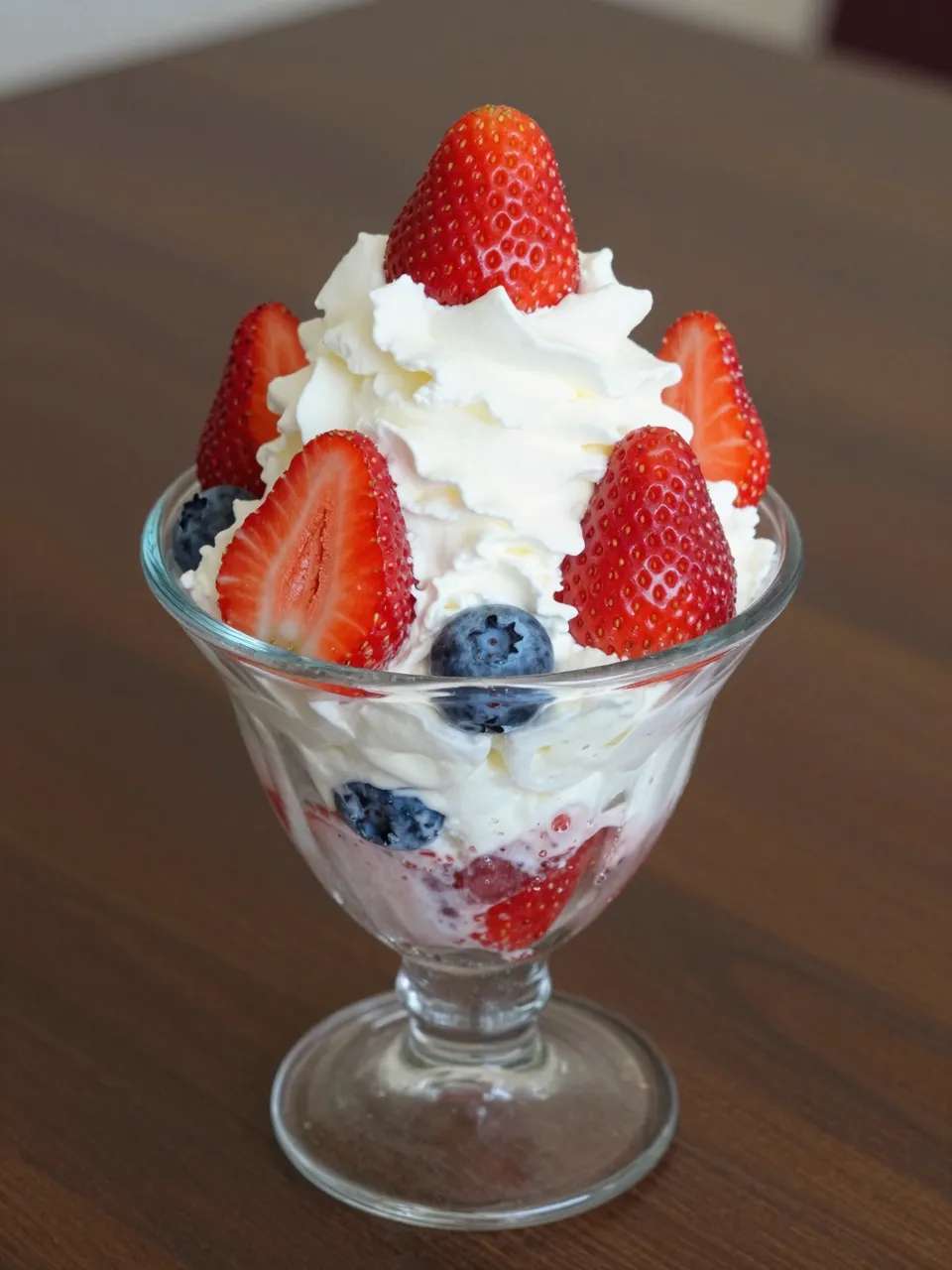 Glass dessert cup filled with whipped cream, fresh strawberries and blueberries resting on a wooden table