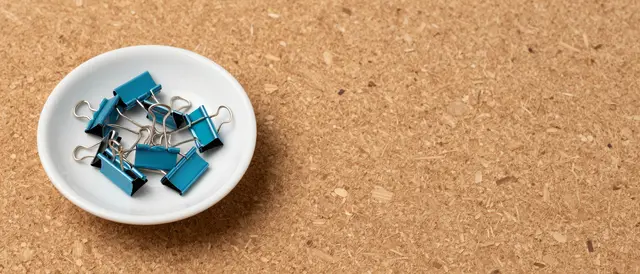 A top-down view of blue binder clips in a small white ceramic dish on a cork surface with empty copy space to the right.