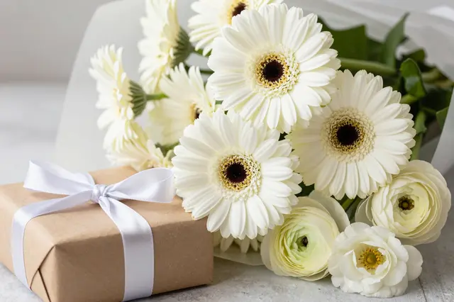 White gerbera daisy and ranunculus bouquet sits beside a kraft wrapped gift with a white satin ribbon on a light surface.