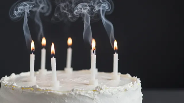 Frosted white birthday cake holds lit candles with curling smoke against a dark background