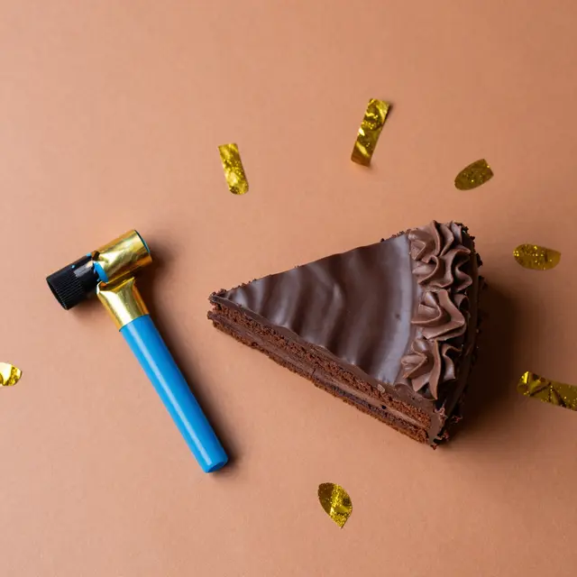 Top-down view of a chocolate cake slice, party blower and gold confetti on a terracotta backdrop with copy space