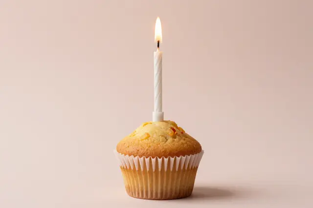 A single cupcake with a lit white birthday candle sits against a soft pastel pink background.