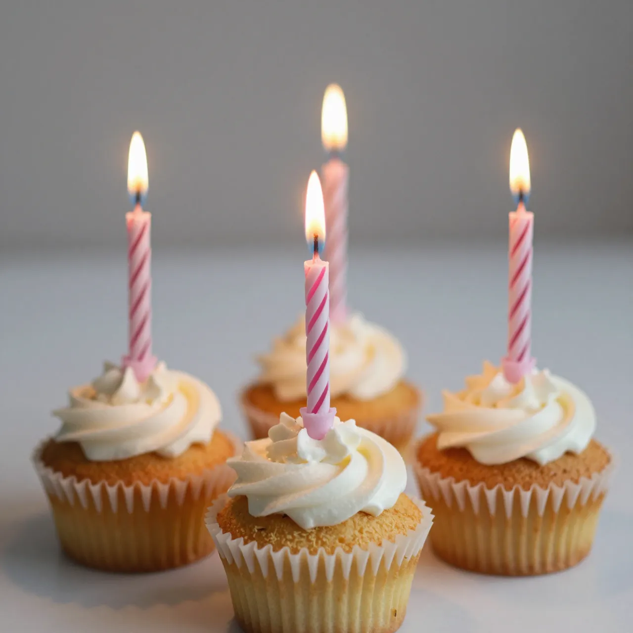 Four vanilla cupcakes with swirled frosting and lit striped birthday candles arranged on a light neutral surface