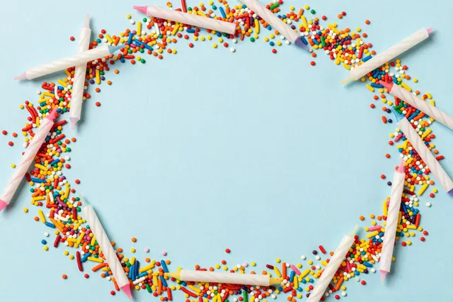 Top view of colorful sprinkles and birthday candles arranged in a circle around empty pale blue space