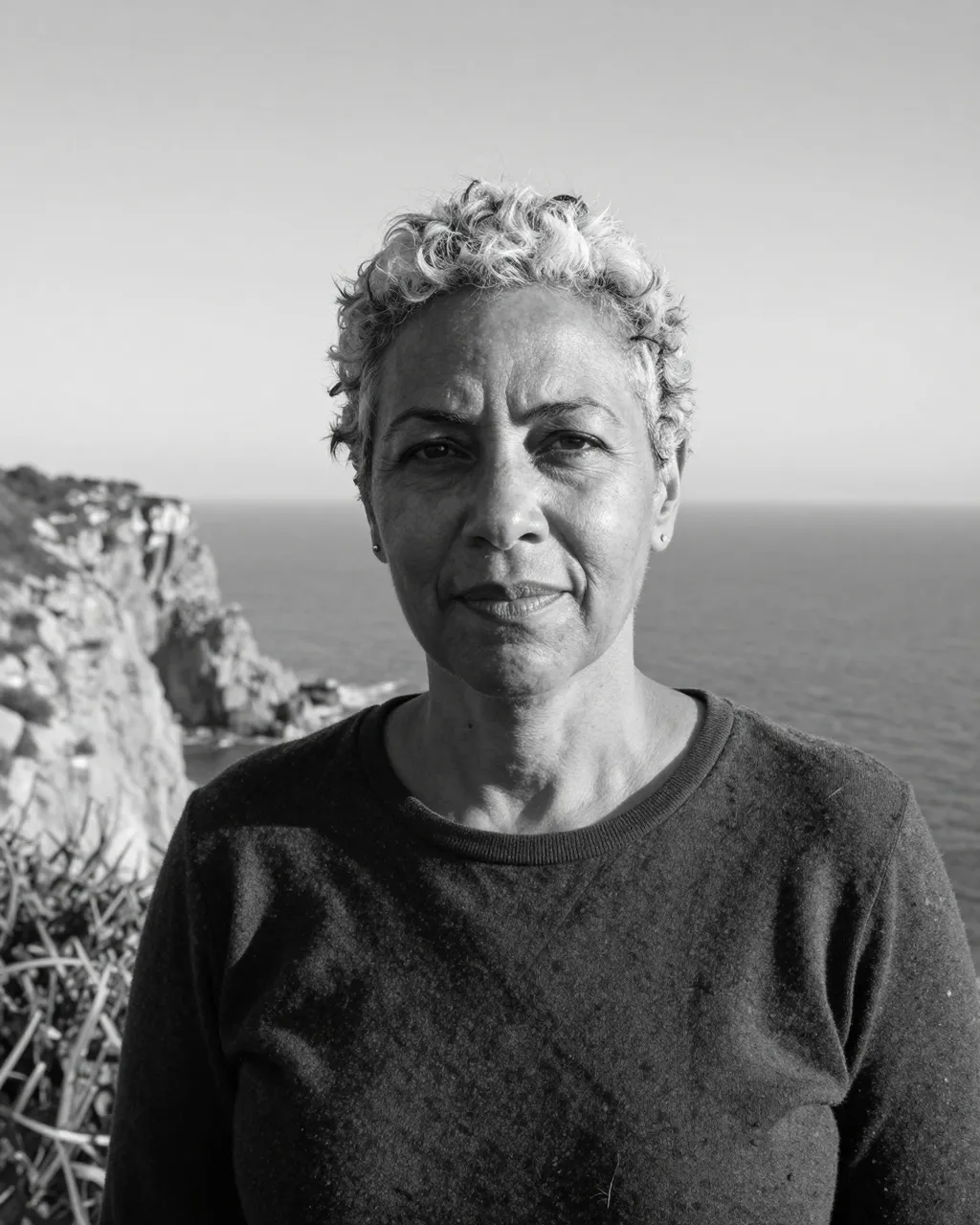 A mixed-race woman in her fifties with cropped gray hair stands on a windy sea cliff in black and white.