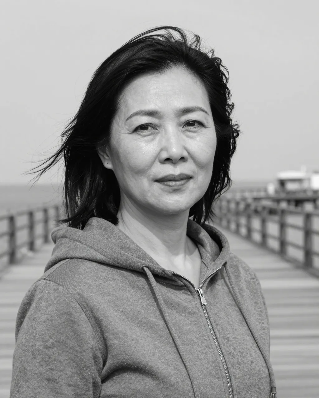 Middle-aged East Asian woman with wind-blown hair stands on a wooden pier in black and white.
