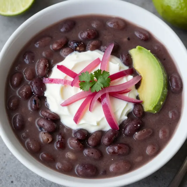 Overhead view of black bean soup topped with sour cream, pickled onion, avocado and cilantro next to lime wedges