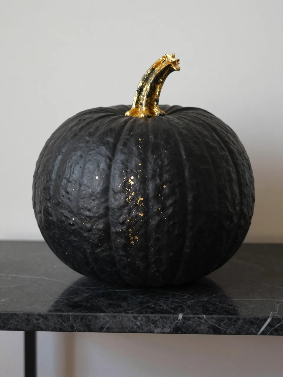 A matte black pumpkin with a gilded stem and gold flecks rests on a dark marble console table.