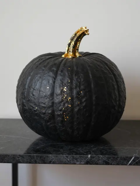 A matte black pumpkin with a gilded stem and gold flecks rests on a dark marble console table.