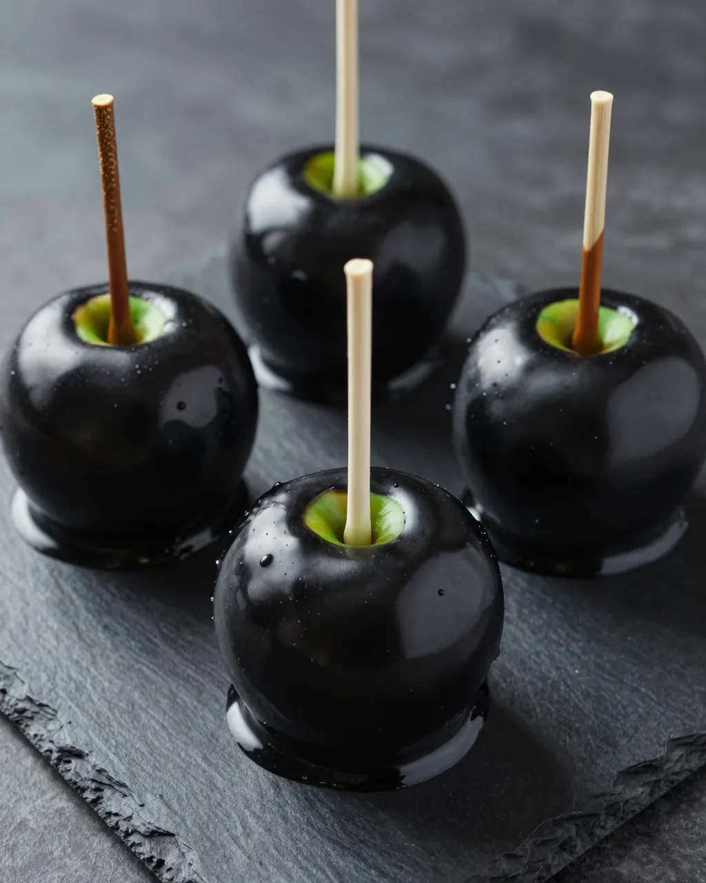 Four glossy black candy apples on a dark slate serving board
