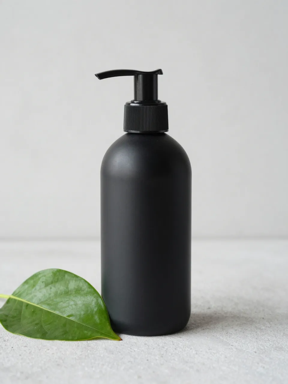 Blank matte black pump bottle next to a green leaf on a light gray concrete surface