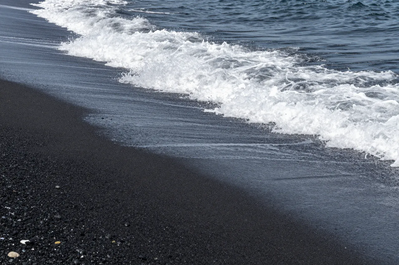 White foamy ocean waves lap onto a dark volcanic black sand shoreline.