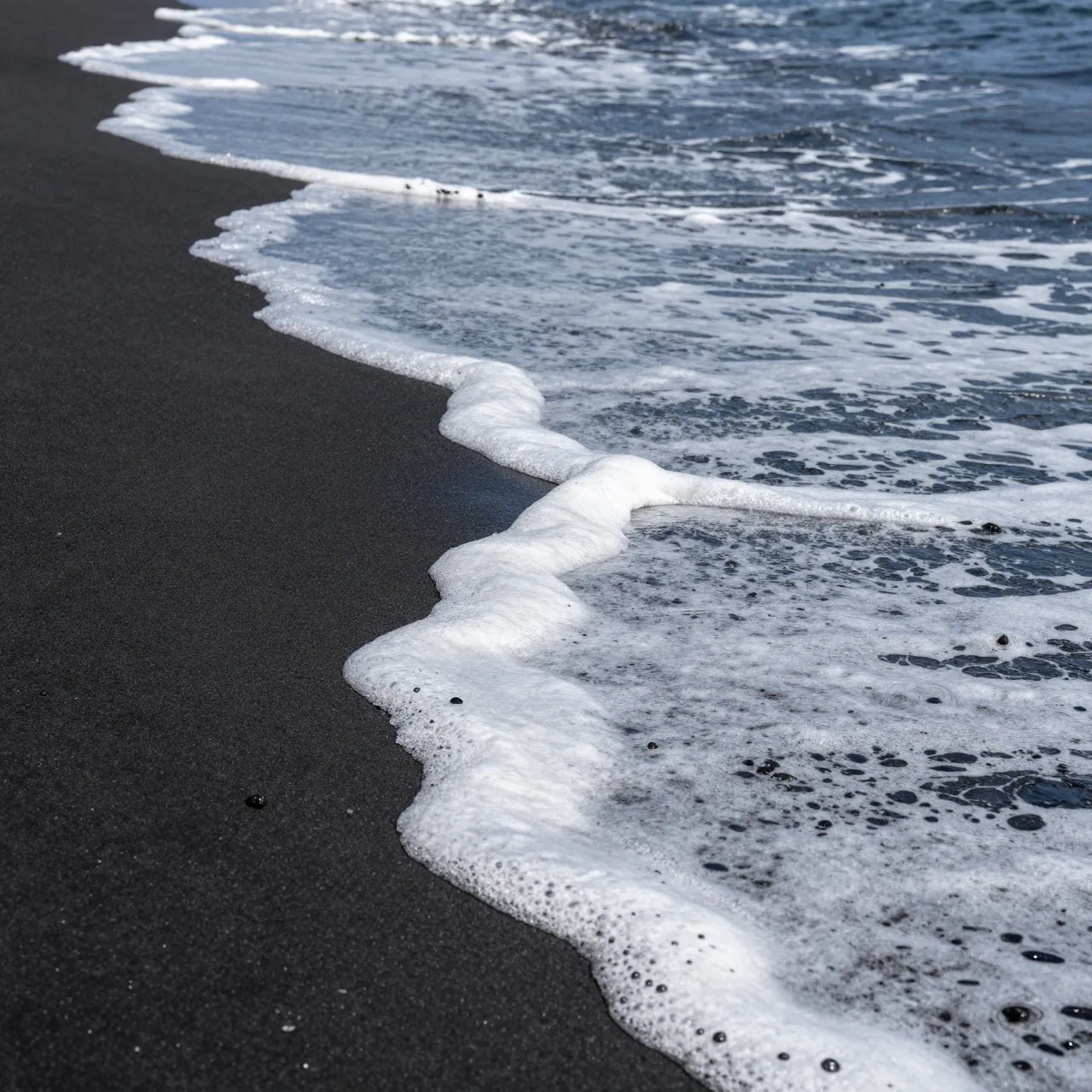 Gentle ocean waves with white foam lap onto a dark black sand beach from a high angle view