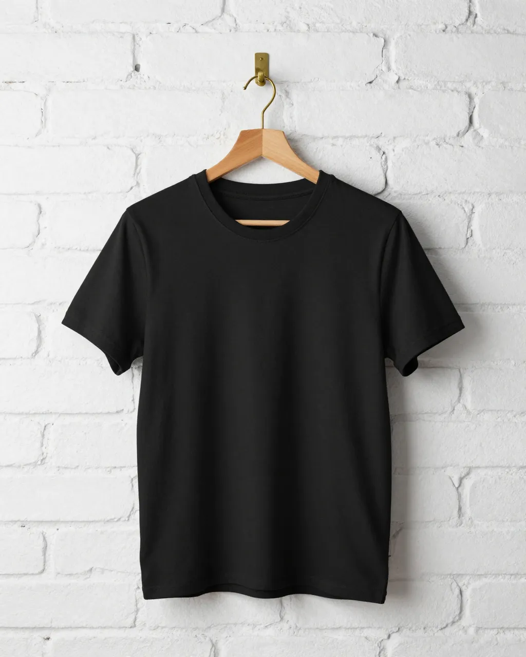 A plain black t-shirt hangs on a wooden hanger attached to a brass hook on whitewashed brick.