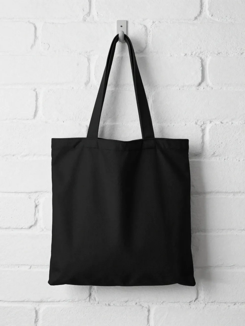 Plain black canvas tote bag hanging from a wall hook against a white painted brick wall