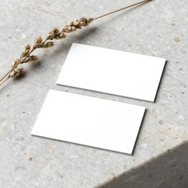 Two blank white business cards rest on a speckled stone slab with a dried grass sprig beside them.