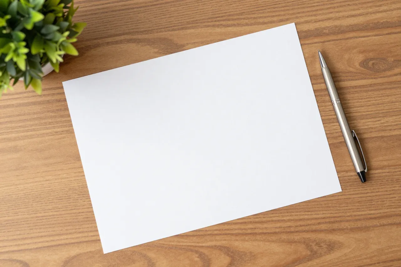 Top-down view of a blank white paper, metal pen and small potted plant resting on a wooden desk