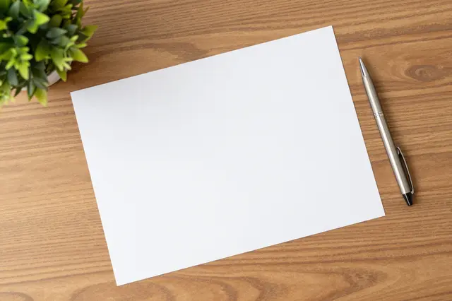 Top-down view of a blank white paper, metal pen and small potted plant resting on a wooden desk