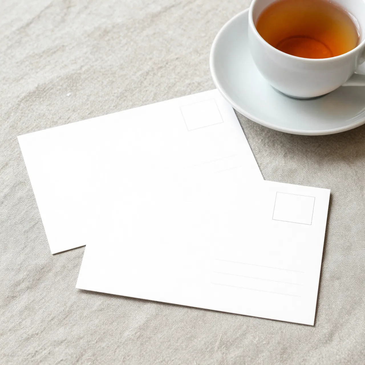Two blank white postcards rest on a linen tablecloth beside a white teacup of tea on a matching saucer