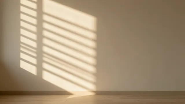 Soft golden window blind shadows fall across a plain cream interior wall and wooden floor with empty copy space.