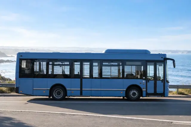 Side view of a plain blue modern bus parked on an asphalt road next to a calm sea on a bright clear morning