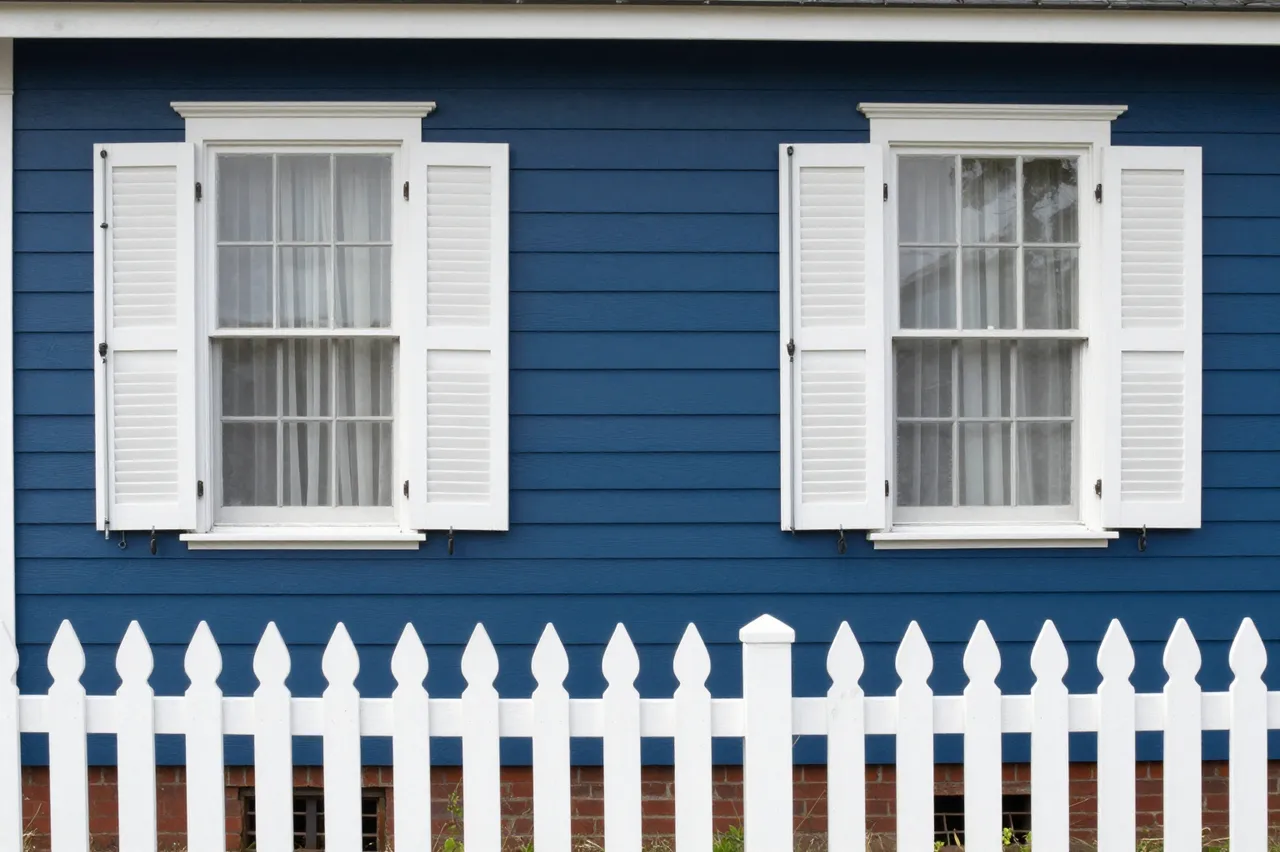 Blue clapboard house wall with white shuttered windows and a white picket fence in the foreground