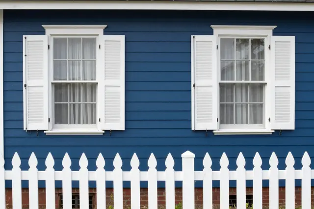 Blue clapboard house wall with white shuttered windows and a white picket fence in the foreground