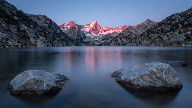 Long exposure view of smooth boulders in a calm mountain lake with pink alpenglow on distant peaks