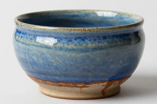 A hand thrown stoneware bowl with reactive blue and rust drip glaze sits against a plain off-white background