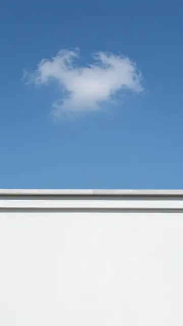 A single wispy cloud floats in a clear bright blue sky above the top edge of a smooth white exterior wall.