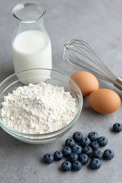 Pancake batter ingredients arranged on a smooth gray countertop: flour, milk, eggs, a whisk, and fresh blueberries