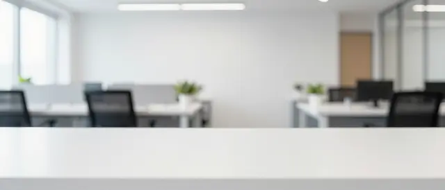 An empty white desk foreground sits before a softly blurred bright modern open office.