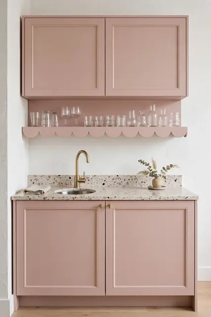 A compact kitchenette with blush cabinets, terrazzo counter, scalloped glassware shelf and brass faucet against a white wall.