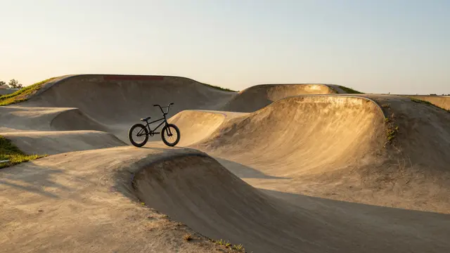 A black BMX bike rests on rolling dirt pump track jumps during warm golden hour light