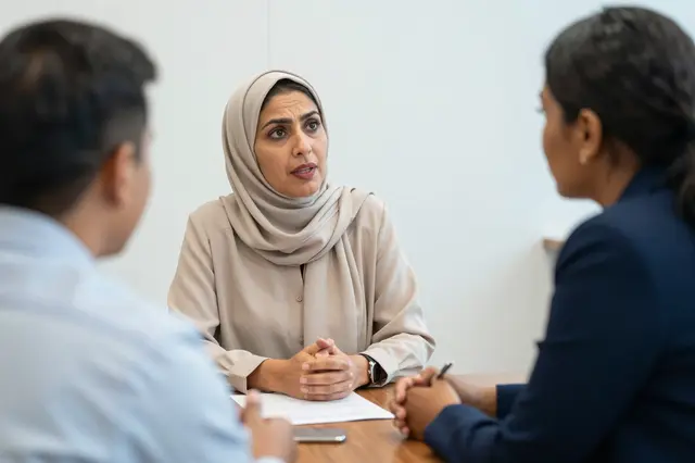 A Middle Eastern woman leads a discussion with two colleagues at a wooden boardroom table.