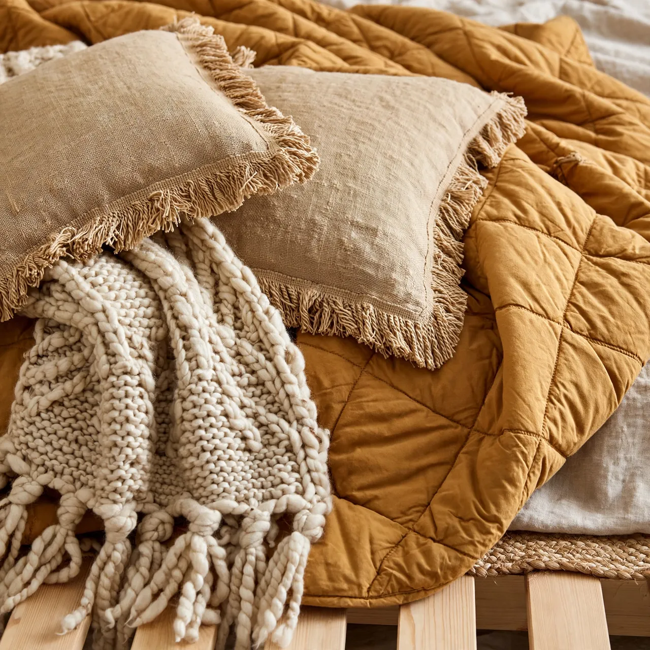Close-up of a platform bed with an ochre quilt, fringed linen pillows and a chunky knit throw